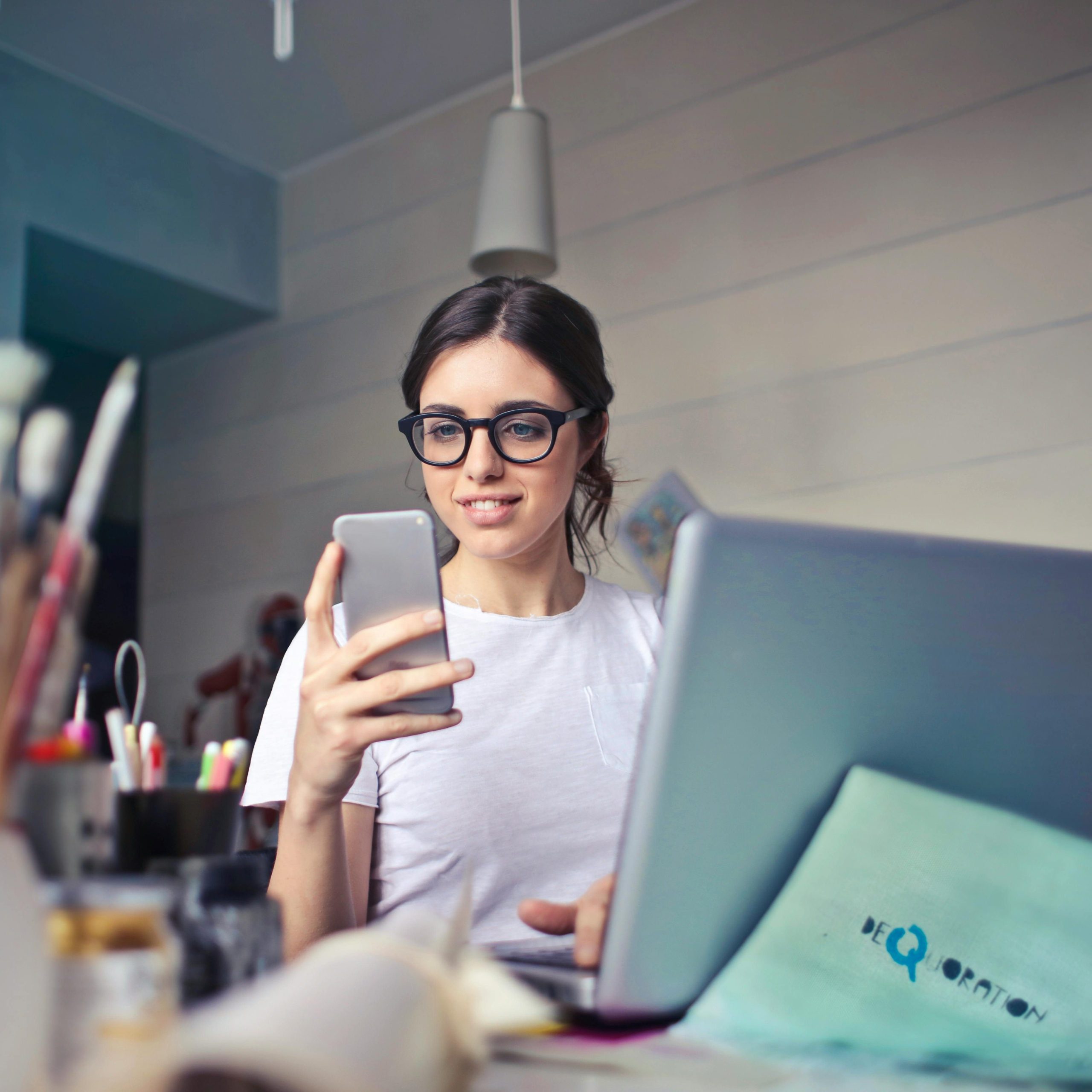 A woman using her phone at a desk, surrounded by art supplies and a laptop, in a creative workspace.